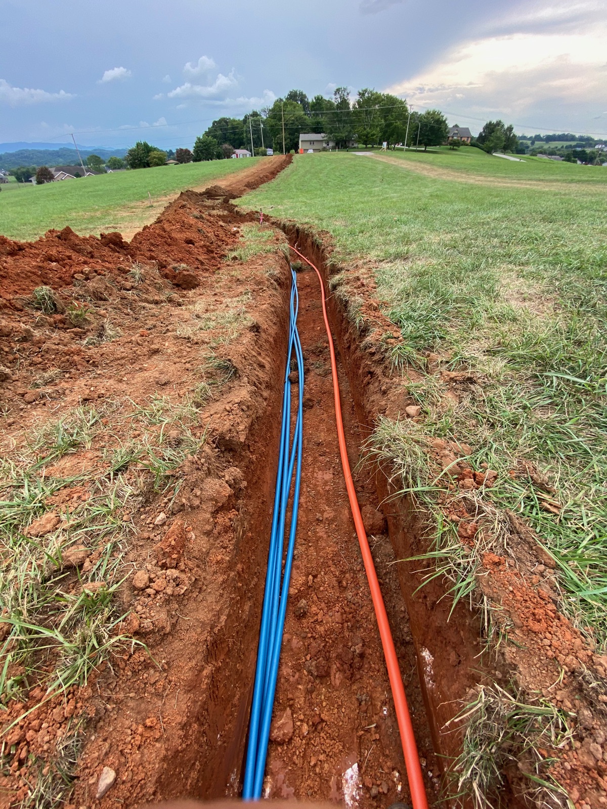 Open trench with blue water line and orange electrical conduit running across pasture